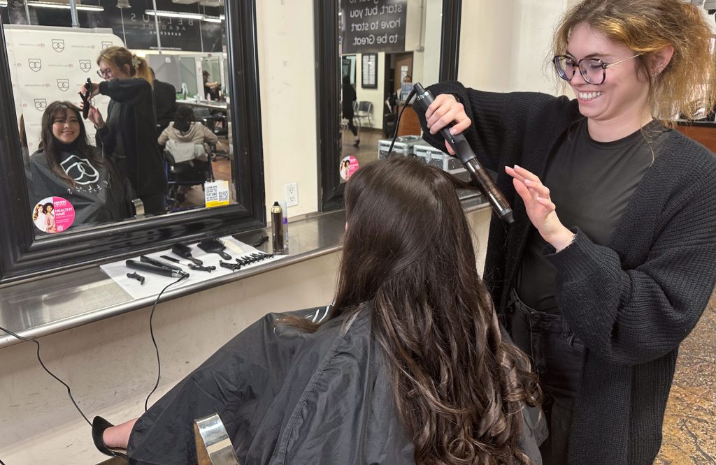 A smiling Bellus Academy Cosmetology student in glasses uses a curling iron on a client's long, dark brown hair. The client, wearing a black cape, smiles in the mirror at a salon station set with brushes, clips, and styling tools.