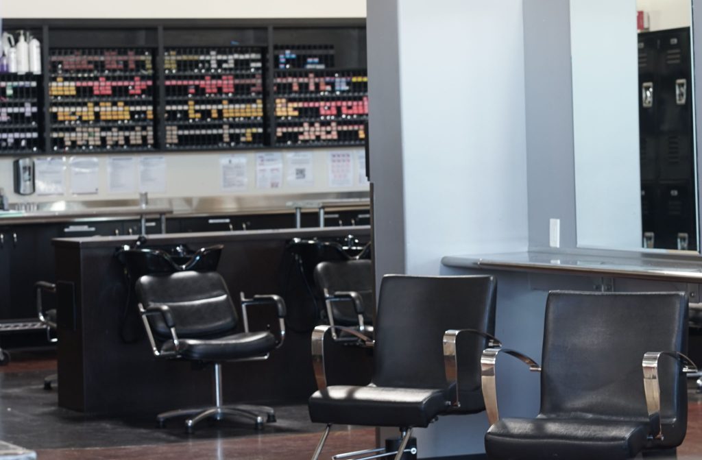 Black leather styling chairs with chrome arms sit in front of a row of shampoo bowls and backwash chairs. Behind them, a wall of shelving holds hair color tubes organized by shade above a stainless steel color bar.