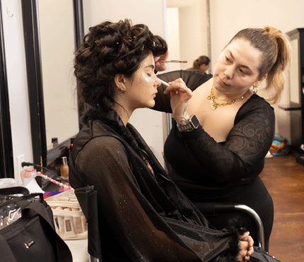 A Bellus Academy student applies makeup with a brush to a model whose eyes are closed and who wears under-eye patches. The model's dark hair is styled in a sculpted, curly updo, and she wears a sheer black robe with feather-trimmed cuffs. Other students work at stations in the background.
