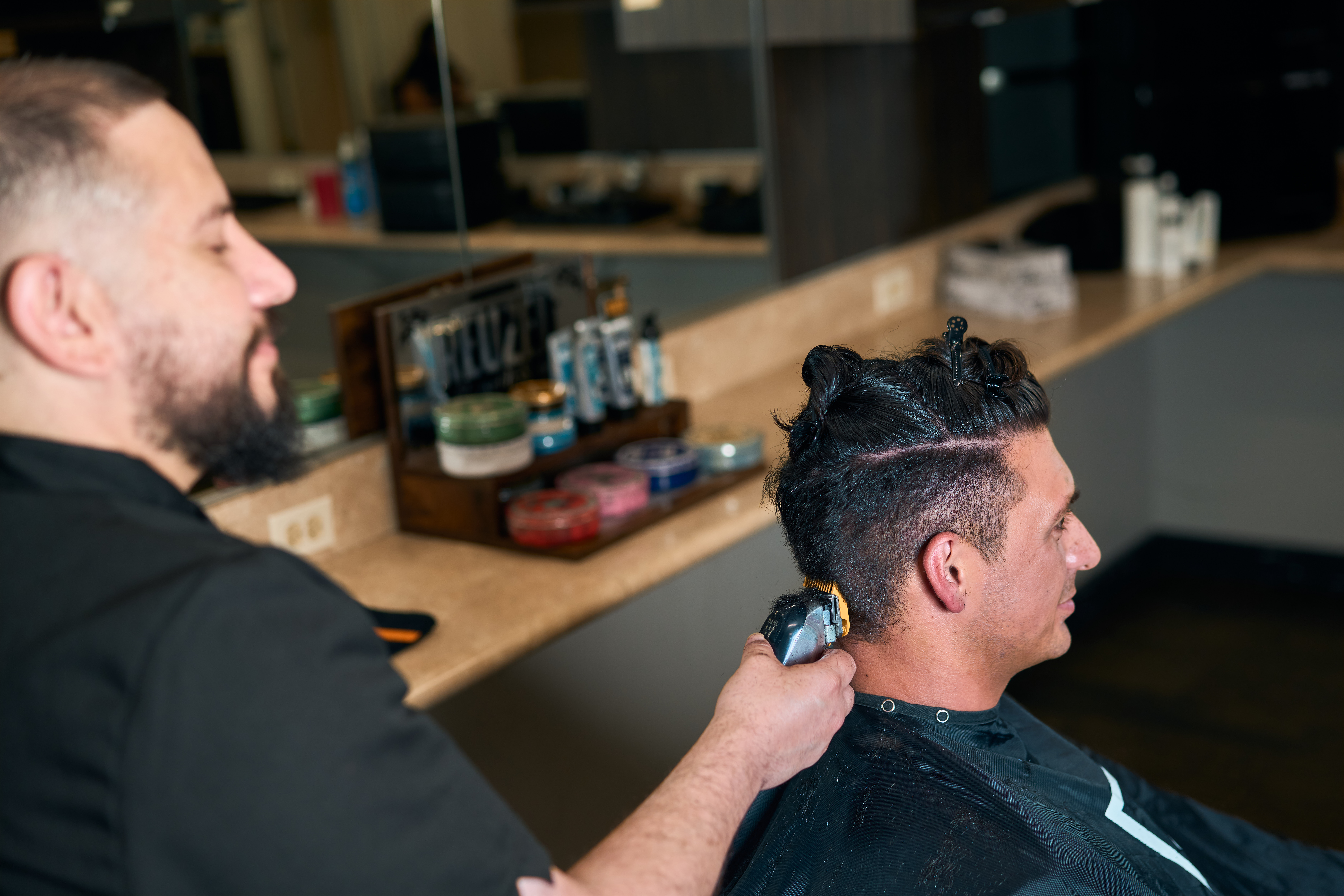 Adrian De La Parra tapers the side of a client’s haircut with clippers at Bellus Academy in Poway, with Reuzel products on the counter.